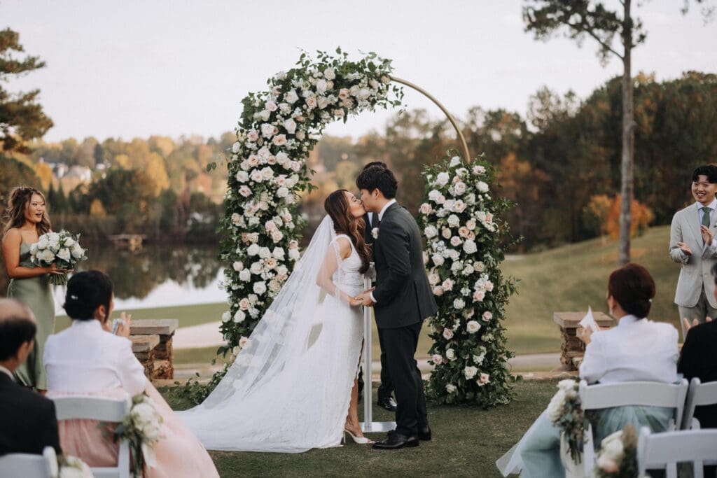 Bride and groom kissing beneath large crescent floral arch during outdoor ceremony at TPC Sugarloaf