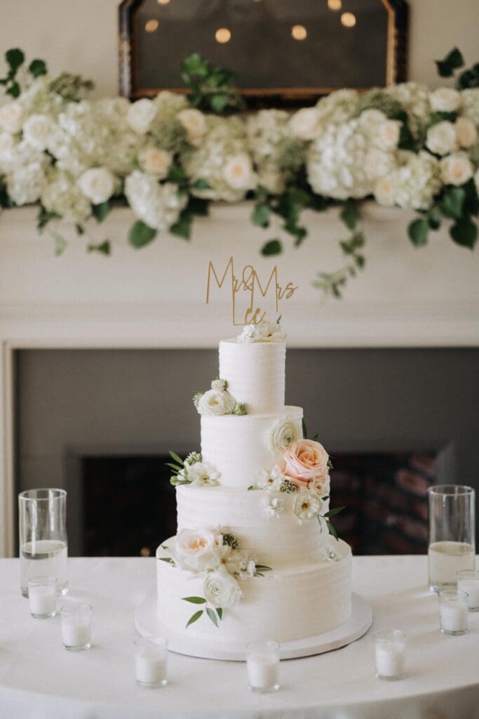 Elegant white wedding cake with blush roses and hydrangea displayed in front of floral fireplace mantle at TPC Sugarloaf