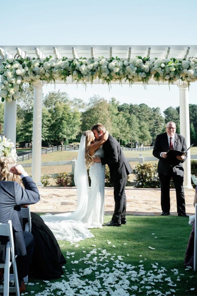 Bride and groom sharing their first kiss beneath a lush white floral pergola at Greenfield Farm.