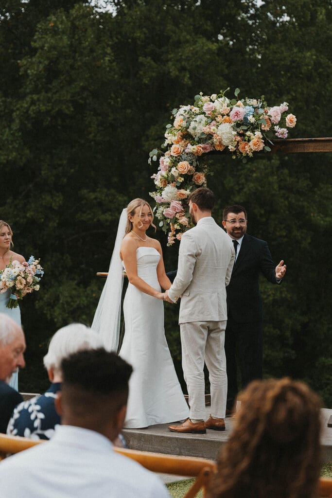 Bride and groom standing under floral ceremony arbor at Meadows at Mossy Creek with mountain backdrop and garden style wedding flowers