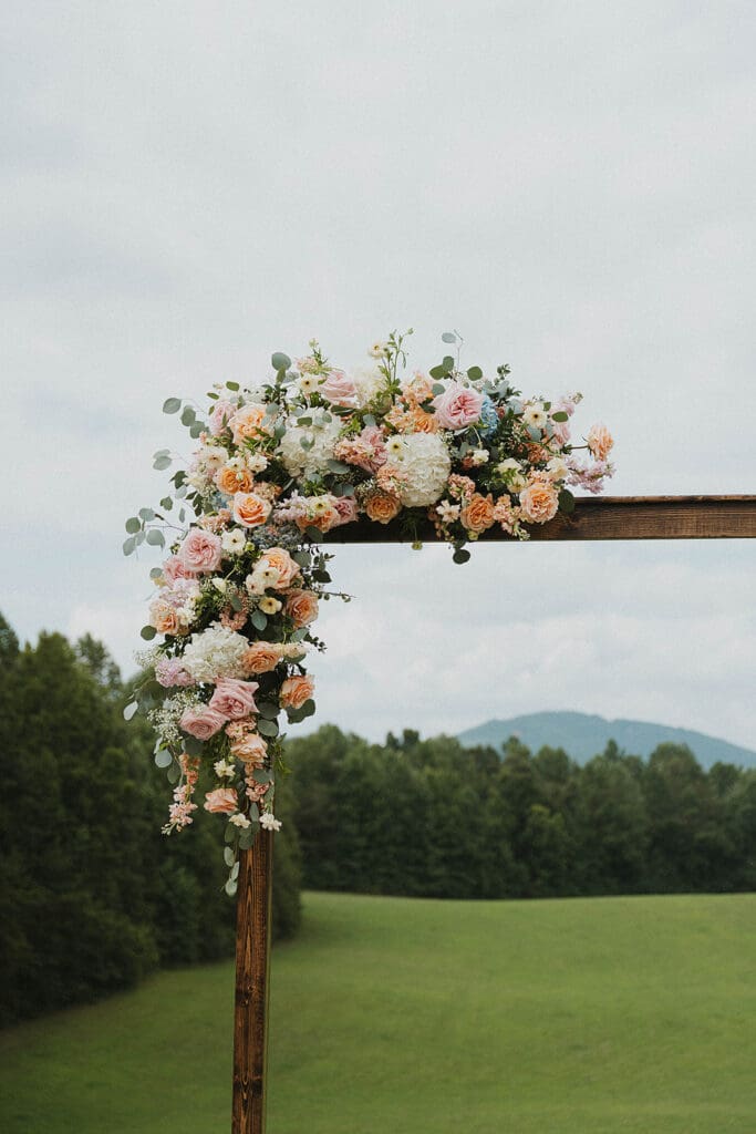 Asymmetrical ceremony arch flowers with peach roses, hydrangea, and pastel garden blooms at Meadows at Mossy Creek wedding in Cleveland Georgia