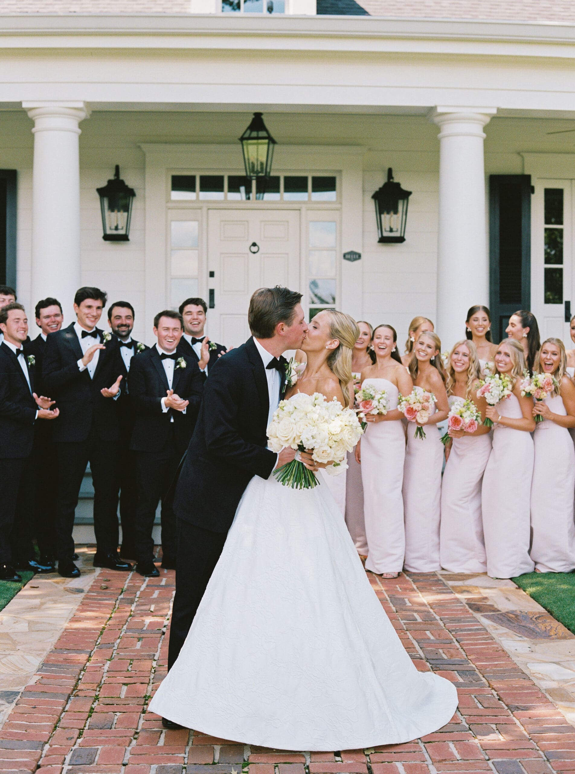 Bride and groom kissing with wedding party outside Greenfield Farms venue