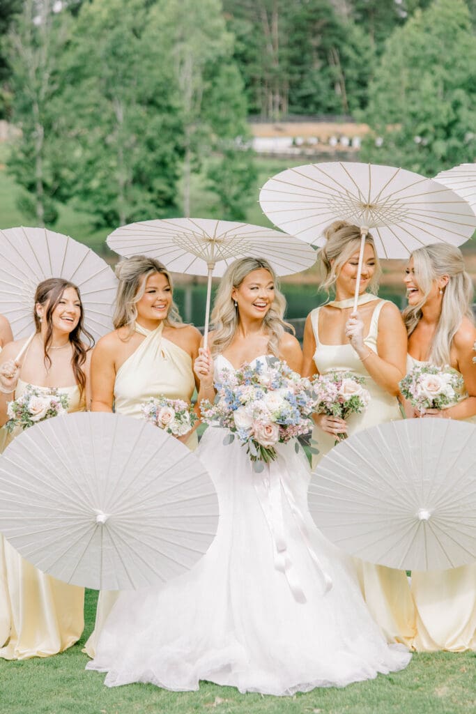 Bride and bridesmaids carrying white parasols and pastel bouquets during summer wedding portraits at The Revere in Dawsonville Georgia.