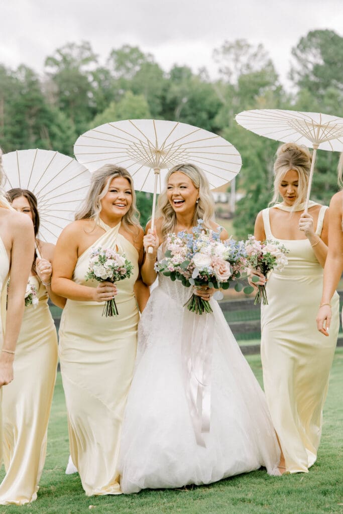 Bride and bridesmaids in soft yellow dresses holding pastel bouquets and white parasols during summer wedding portraits at The Revere in Dawsonville Georgia.