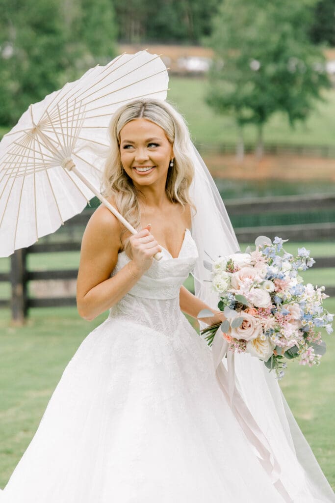 Bride holding a pastel garden style bouquet and white parasol during summer wedding portraits at The Revere wedding venue in Dawsonville Georgia.