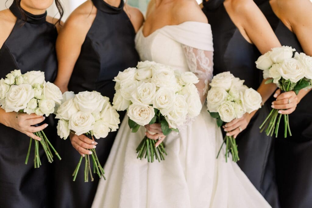 Bride with bridesmaids in black dresses holding white rose bouquets at TPC Sugarloaf wedding
