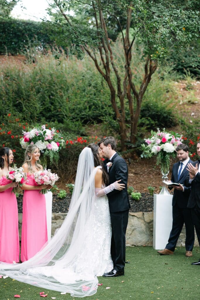 Bride and groom sharing their first kiss surrounded by pink and white ceremony flowers during an outdoor wedding at Country Club of the South.