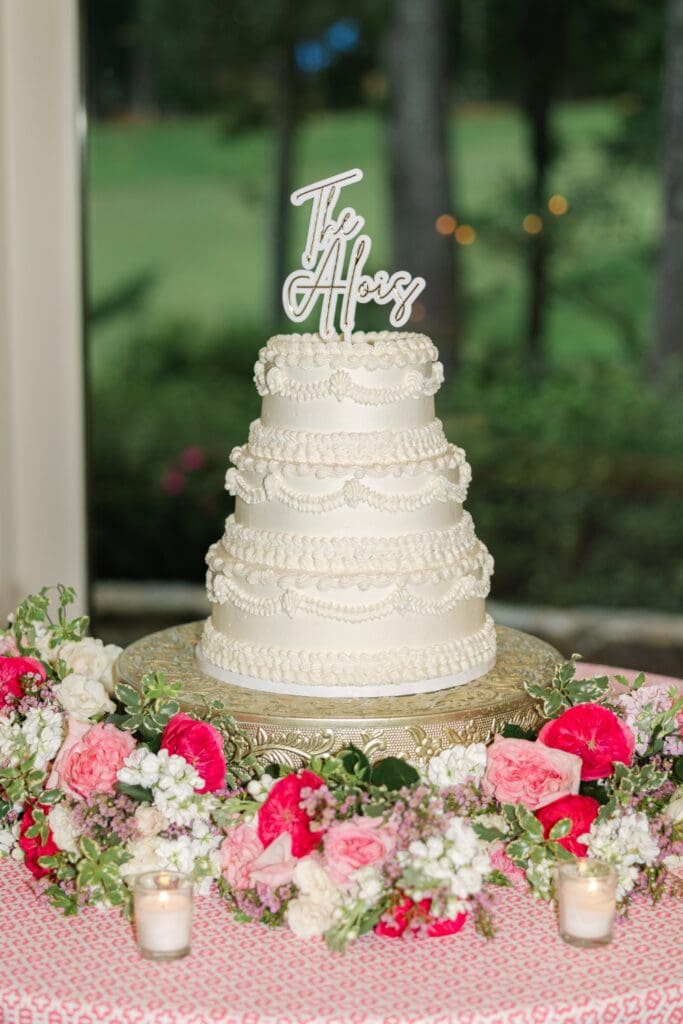 Elegant white wedding cake surrounded by pink and white floral meadow arrangements with roses, hydrangeas, and candles at Country Club of the South wedding reception.