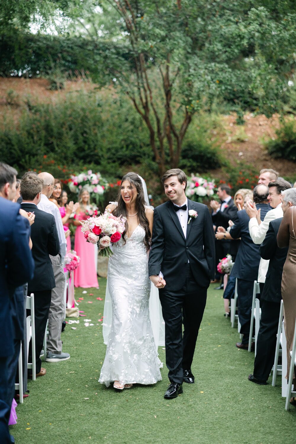 Newly married couple walking down the ceremony aisle surrounded by guests and vibrant pink wedding flowers.