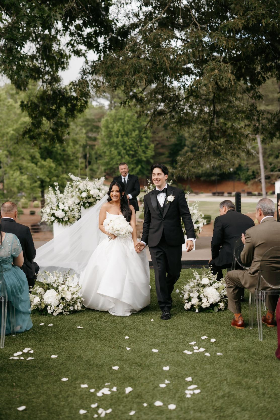 Newlyweds walking down an outdoor aisle lined with white wedding flowers